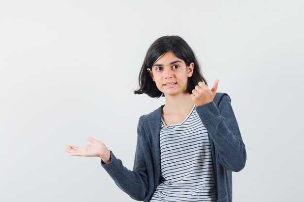 Kleines Mädchen im T-Shirt, Jacke spreizt Handfläche beiseite, zeigt Daumen hoch und sieht selbstbewusst aus