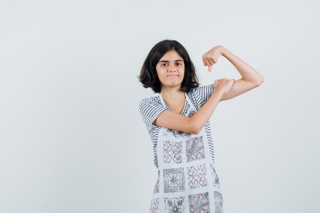 Kleines Mädchen im T-Shirt, Schürze, die Armmuskeln zeigt und selbstbewusst aussieht,