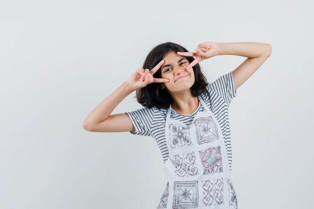 Kleines Mädchen im T-Shirt, Schürze, die V-Zeichen nahe Augen zeigt und fröhlich aussieht,