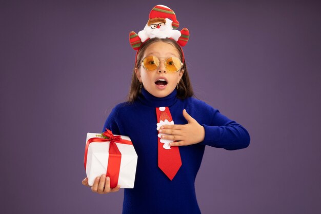 Kleines Mädchen in blauem Rollkragenpullover mit roter Krawatte und lustigem Weihnachtsrand auf dem Kopf, das ein Geschenk glücklich und positiv mit der Hand auf der Brust hält und sich dankbar fühlt, über lila Wand zu stehen