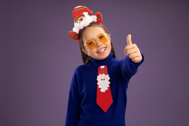 Kleines Mädchen in blauem Rollkragenpullover mit roter Krawatte und lustigem Weihnachtsrand auf dem Kopf glücklich und positiv lächelnd mit Daumen nach oben stehend über rosa Wand