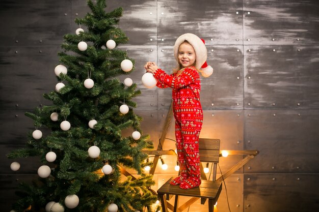 Kleines Mädchen in den Pyjamas durch den Weihnachtsbaum auf einem Holzstuhl