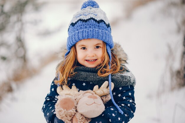 Kleines Mädchen in einem blauen Hut, der in einem Winterwald spielt