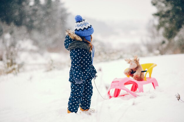 Kleines Mädchen in einem blauen Hut, der in einem Winterwald spielt