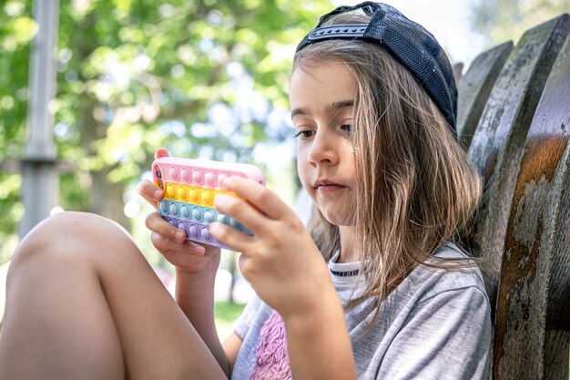 Kleines Mädchen in einer Mütze mit einem Smartphone in einer Hülle im Stil von Spielzeug Anti-Stress-Pop it.