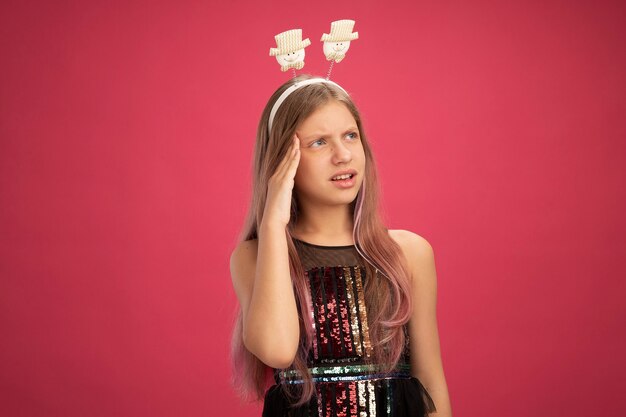 Kleines Mädchen in Glitzer-Partykleid und lustigem Stirnband, das verwirrt beiseite schaut, Neujahrsfeier-Feiertagskonzept, das über rosafarbenem Hintergrund steht