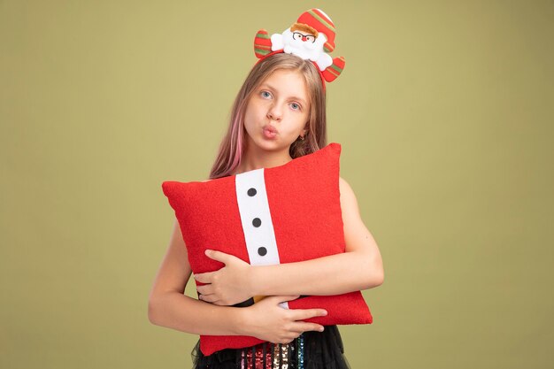 Kleines Mädchen in Glitzer-Partykleid und Stirnband mit Weihnachtsmann, der ein lustiges Kissen hält und die Kamera gelangweilt auf grünem Hintergrund sieht