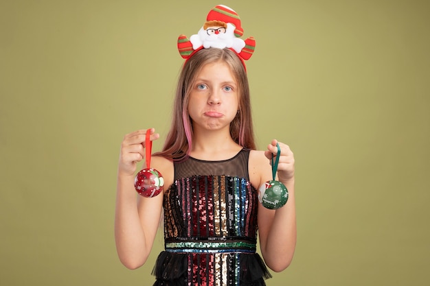 Kleines Mädchen in Glitzer-Partykleid und Stirnband mit Weihnachtsmann, der Weihnachtskugeln hält, die verwirrt und unzufrieden in die Kamera schaut, über grünem Hintergrund stehend