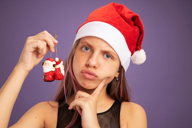 Kleines Mädchen in Glitzer-Partykleid und Weihnachtsmütze mit Weihnachtsspielzeug, das mit ernstem Gesicht auf lila Hintergrund in die Kamera schaut