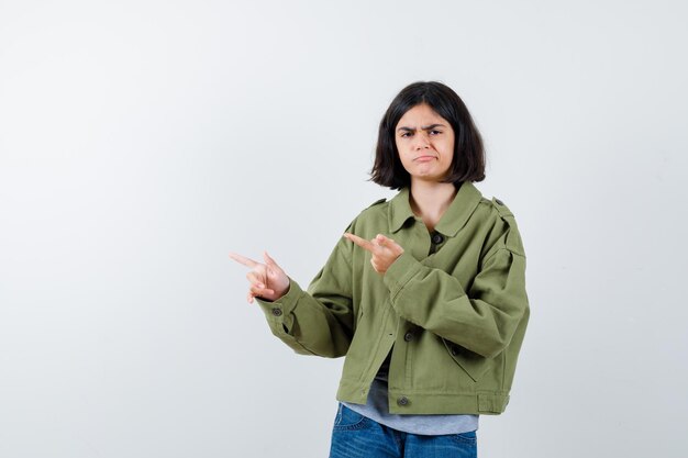 Kleines Mädchen in Mantel, T-Shirt, Jeans, die auf die linke Seite zeigt und nachdenklich aussieht, Vorderansicht.