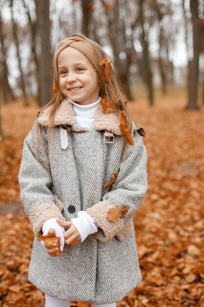 Kleines Mädchen in modischer Kleidung, die im Herbstwald steht Mädchen, das für ein Foto aufwirft Mädchen, das grauen Mantel und ein Stirnband trägt