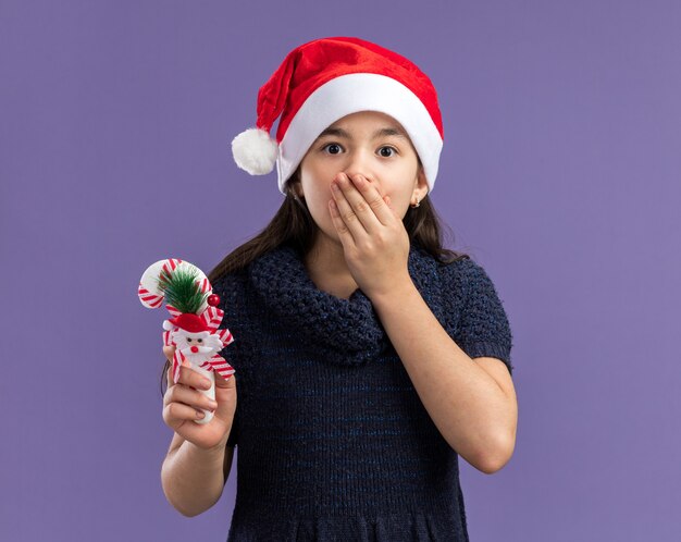 Kleines Mädchen in Strickkleid mit Weihnachtsmütze, das Weihnachtszuckerstangen hält und schockiert ist, um den Mund mit der Hand zu bedecken, die über der lila Wand steht