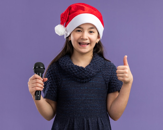 Kleines Mädchen in Strickkleid mit Weihnachtsmütze mit Mikrofon glücklich und positiv, das Daumen hoch steht über lila Wand