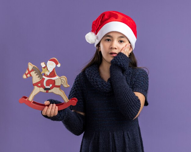 Kleines Mädchen in Strickkleid mit Weihnachtsmütze mit Weihnachtsspielzeug erstaunt und überrascht über lila Wand stehend