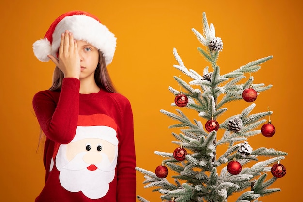 Kleines Mädchen in Weihnachtspullover und Weihnachtsmütze, das die Kamera mit ernstem Gesicht betrachtet, das das Auge mit der Hand bedeckt, die neben einem Weihnachtsbaum über orangefarbenem Hintergrund steht