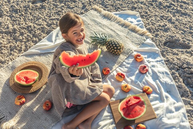 Kleines Mädchen isst Obst, das auf einer Decke am Strand liegt