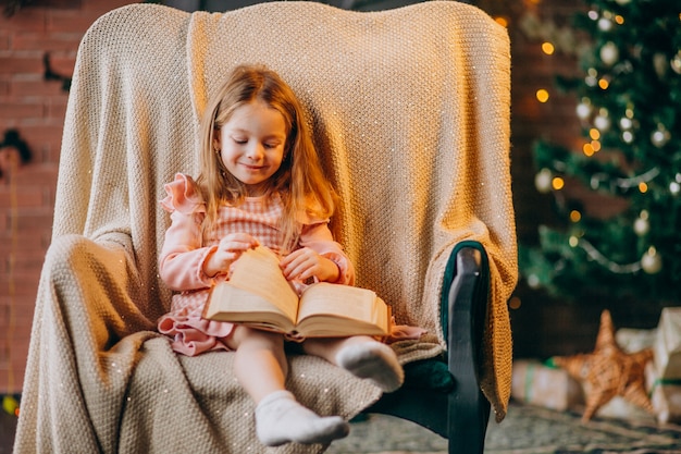 Kleines Mädchen mit dem Buch, das im Stuhl durch Weihnachtsbaum sitzt