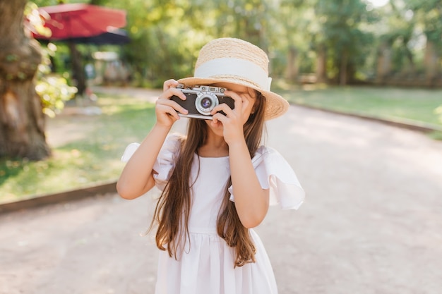 Kleines Mädchen mit dem langen dunklen Haar, das Kamera in den Händen hält, die auf der Gasse im Park stehen. Weibliches Kind im Strohhut mit weißem Band, das Foto der Naturansicht im sonnigen Tag nimmt.