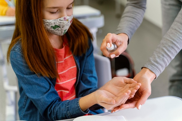 Kleines Mädchen mit der medizinischen Maske, die Händedesinfektionsmittel vom Lehrer erhält