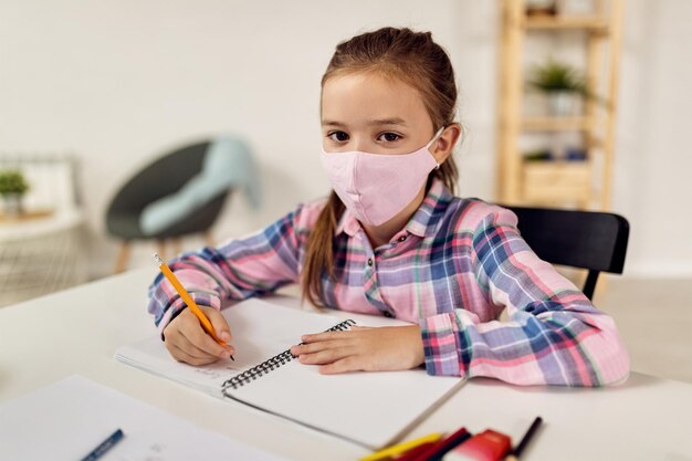 Kleines Mädchen mit Gesichtsmaske, das während der COVID19-Pandemie zu Hause lernt