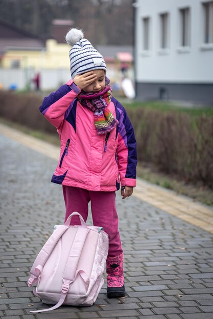 Kleines Mädchen mit Rucksack in Jacke und Hut in der Nähe der Schule