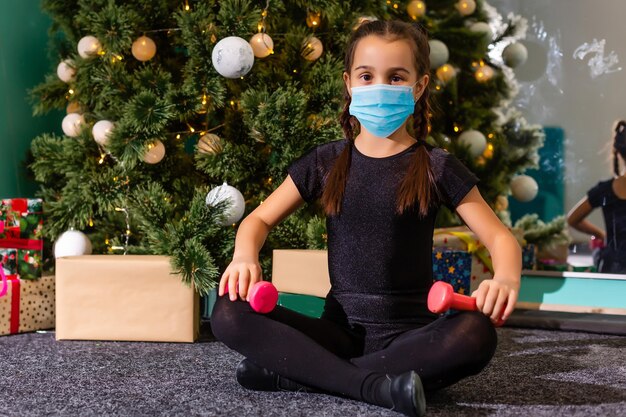 Kleines Mädchen mit Schutzmaske auf Weihnachtsbaumhintergrund. Trauriges Kindergesicht mit OP-Maske. Weihnachtsfeier und soziale Distanzierung während des Covid-19-Pandemiekonzepts.