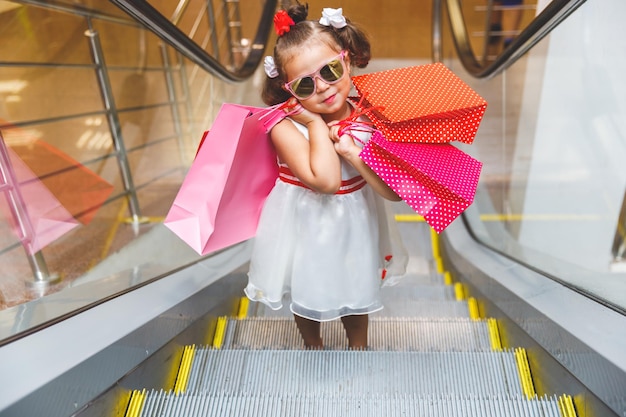 Kleines mädchen mit sonnenbrille auf der rolltreppe im einkaufszentrum mit einkäufen