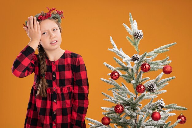 Kleines Mädchen mit Weihnachtskranz im karierten Hemd, das verwirrt mit der Hand auf dem Kopf aussieht, die neben einem Weihnachtsbaum über der orangefarbenen Wand steht?