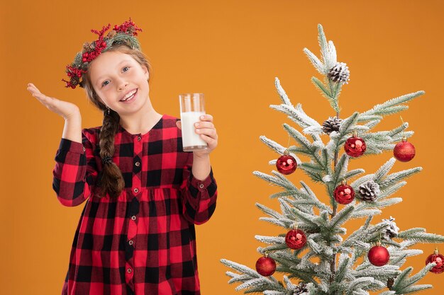 Kleines Mädchen mit Weihnachtskranz in kariertem Hemd, das ein Glas Milch hält, glücklich und positiv lächelnd, fröhlich neben einem Weihnachtsbaum über orangefarbener Wand stehen standing