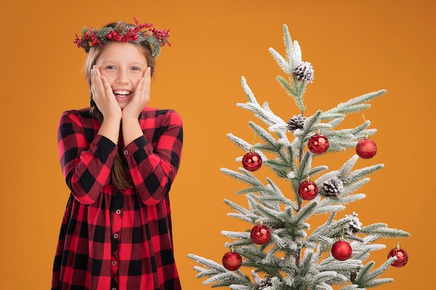 Kleines Mädchen mit Weihnachtskranz in kariertem Hemd erstaunt und überrascht, neben einem Weihnachtsbaum über orangefarbener Wand zu stehen