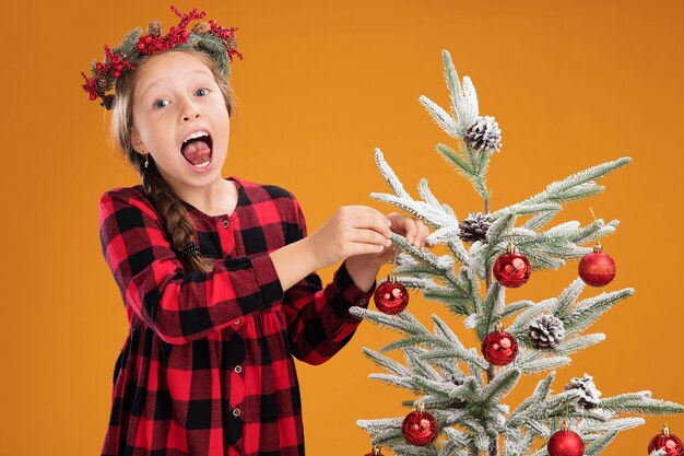 Kleines Mädchen mit Weihnachtskranz in kariertem Kleid, das den Weihnachtsbaum glücklich und freudig schmückt und die Zunge über die orangefarbene Wand streckt