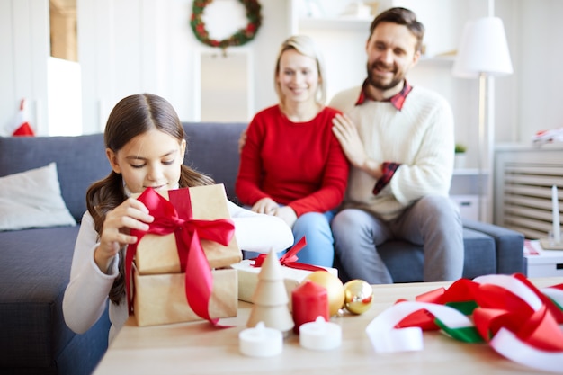 Kleines Mädchen packt das Weihnachtsgeschenk aus, während ihre Eltern sie glücklich ansehen.