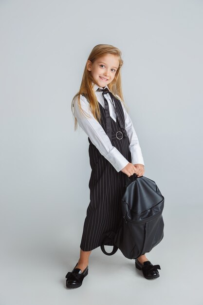 Kleines Mädchen posiert in Schuluniform mit Rucksack auf weißer Wand