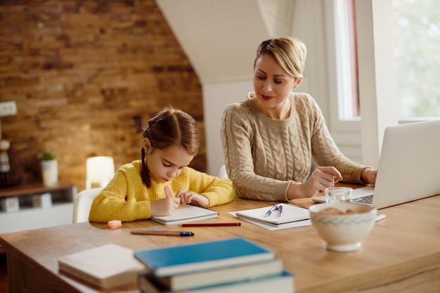 Kleines Mädchen schreibt in Notizbuch, während ihre Mutter zu Hause am Laptop arbeitet