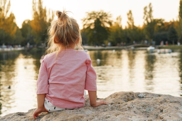 Kleines Mädchen sitzt auf einem riesigen Stein im Park in der Nähe des Sees