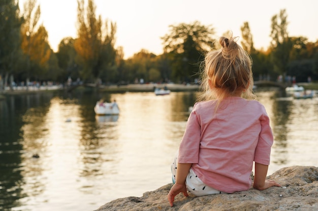 Kleines Mädchen sitzt auf einem riesigen Stein im Park in der Nähe des Sees