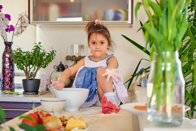 Kleines Mädchen sitzt auf einem Tisch in einer Küche und versucht, Diätbrei zuzubereiten.