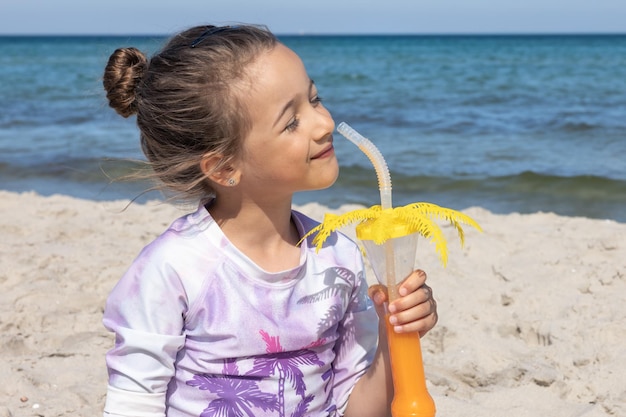 Kleines Mädchen trinkt Saft, der auf dem Sand nahe dem Meer sitzt