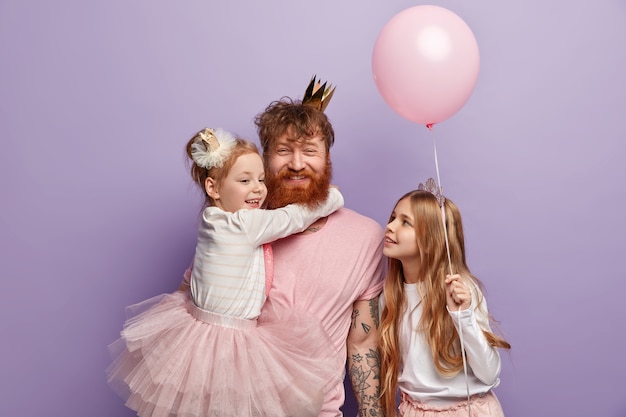 Kleines Mädchen umarmt rothaarigen Vater, der glücklich lächelt, froh, zwei Töchter zu haben, gekleidet in festliche Kleidung, Vatertag feiern, Ballon halten, isoliert über lila Wand. Kinder, Urlaub, Familie
