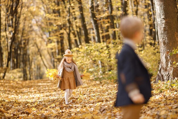 Kleines Mädchen und Junge im Herbstpark. Mädchen, das gelbe Blätter hält und geht. Junge ist auf dem Foto verschwommen.