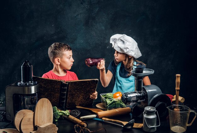 Kleines Mädchen und Junge kochen Gemüse und lesen alte Rezeptbücher.