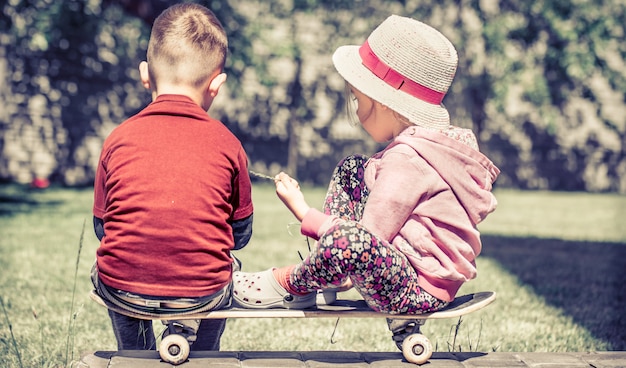 Kleines mädchen und junge spielen auf skateboard, gegen im grünen garten, konzept der kindheitsfreundschaft