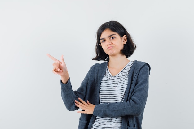 Kleines Mädchen zeigt auf die obere linke Ecke in T-Shirt, Jacke und schaut zögernd.