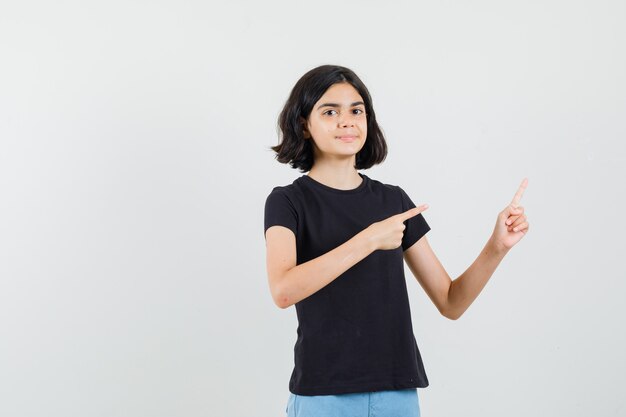 Kleines Mädchen zeigt auf die obere rechte Ecke in schwarzem T-Shirt, Shorts und selbstbewusst aussehend, Vorderansicht.