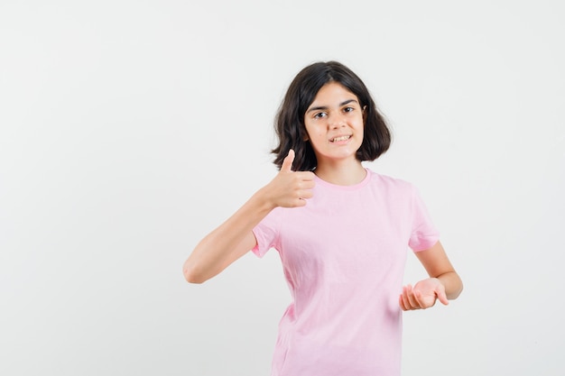 Kleines Mädchen zeigt Daumen hoch, hält Handfläche offen in rosa T-Shirt und sieht fröhlich aus. Vorderansicht.