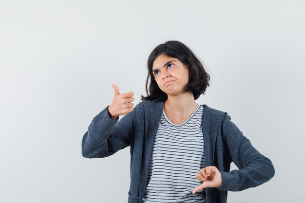 Kleines Mädchen zeigt Daumen hoch und runter in T-Shirt, Jacke und zögert.