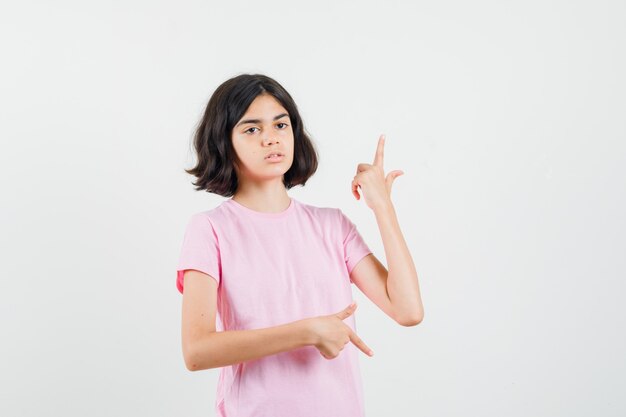 Kleines Mädchen zeigt Finger auf und ab in rosa T-Shirt und schaut zögernd, Vorderansicht.