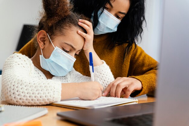 Kleines Mädchen zu Hause, das medizinische Maske während der Online-Schule mit großer Schwester trägt