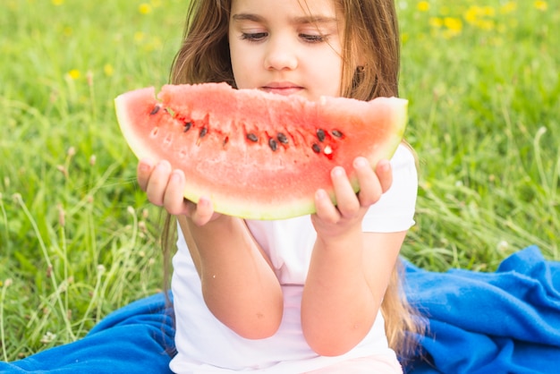Kleines nettes Mädchen, das rote Scheibe der Wassermelone im Park hält