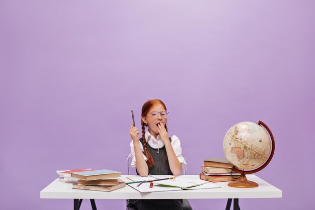 Kleines rothaariges Mädchen mit Zöpfen in Brille und grauem Sommerkleid, das gähnt und mit Büchern und Globus auf violettem Hintergrund am Schreibtisch sitzt